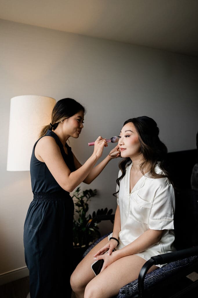 East Asian bride sitting in a makeup chair with makeup artist