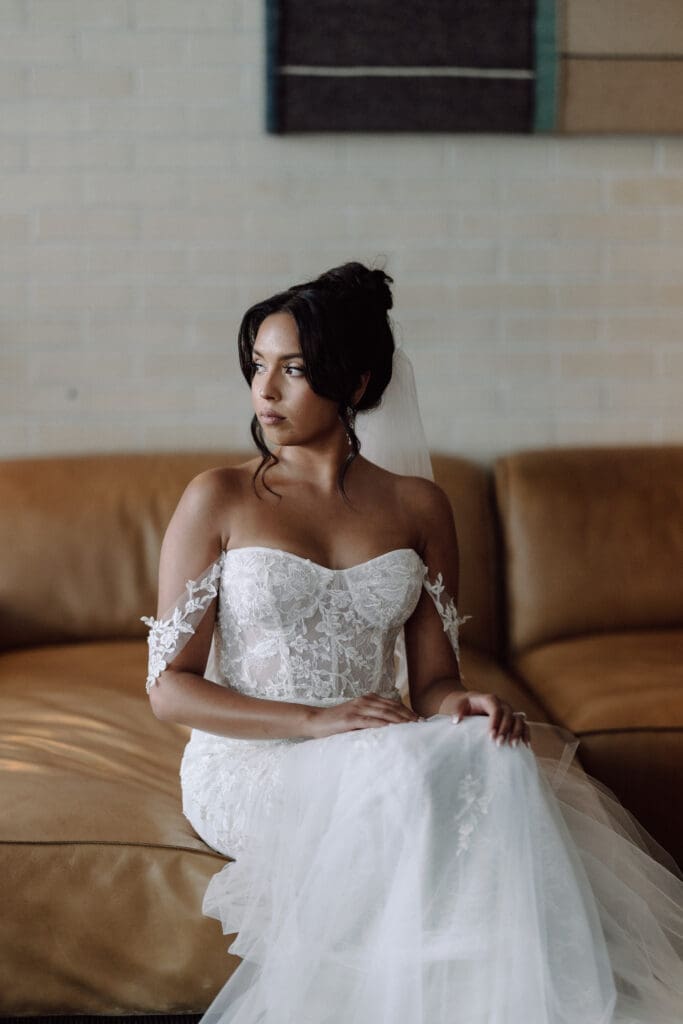 Bride wearing off shoulder wedding gown sitting on a couch.